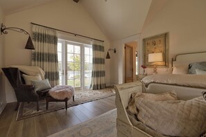 Ein Schlafzimmer mit einem großen Bett, einem Sessel, einem kleinen Tisch und einem Fenster mit Vorhängen.
