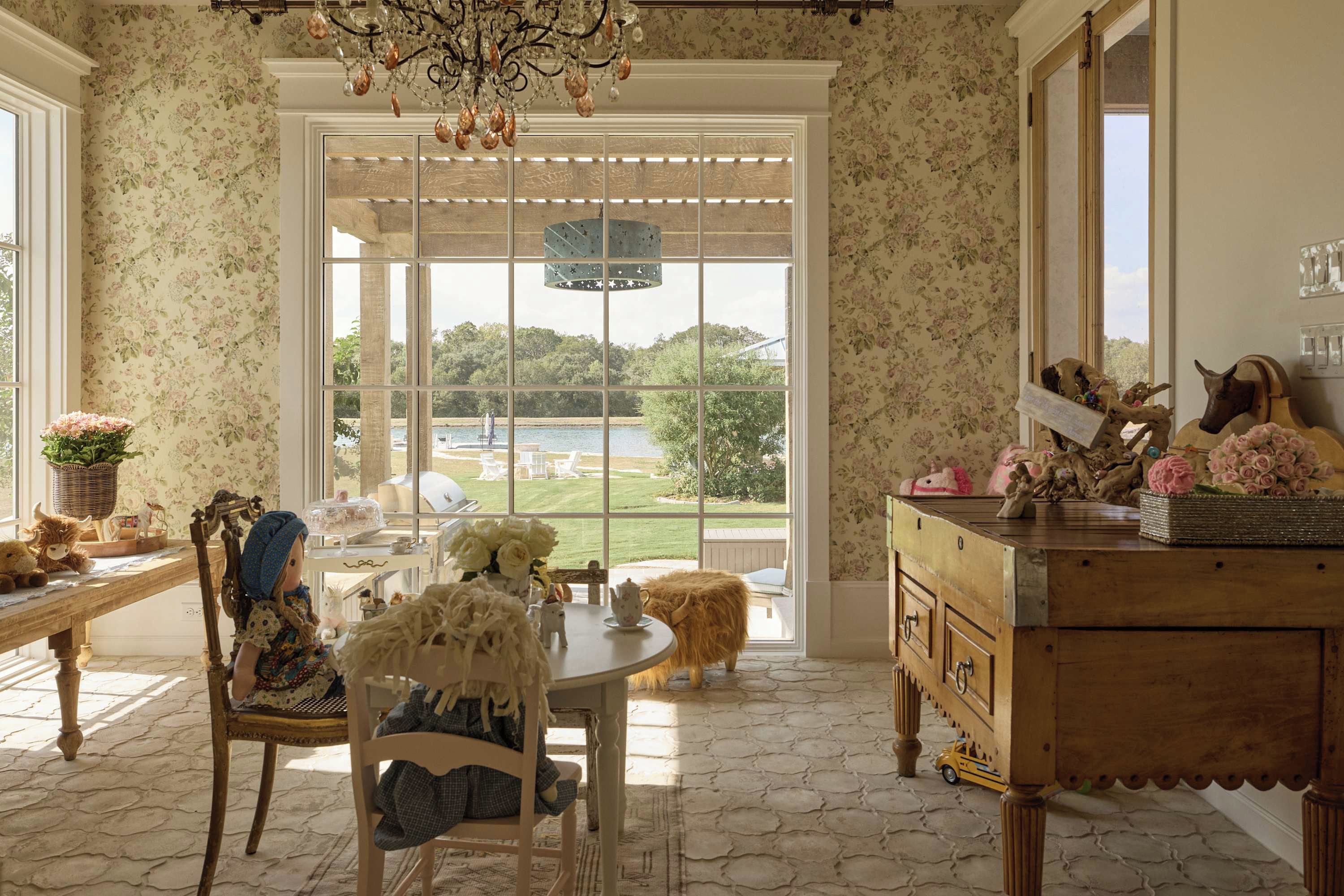 A room with a large window, a chandelier, a wooden table, and a cabinet with decorative items.