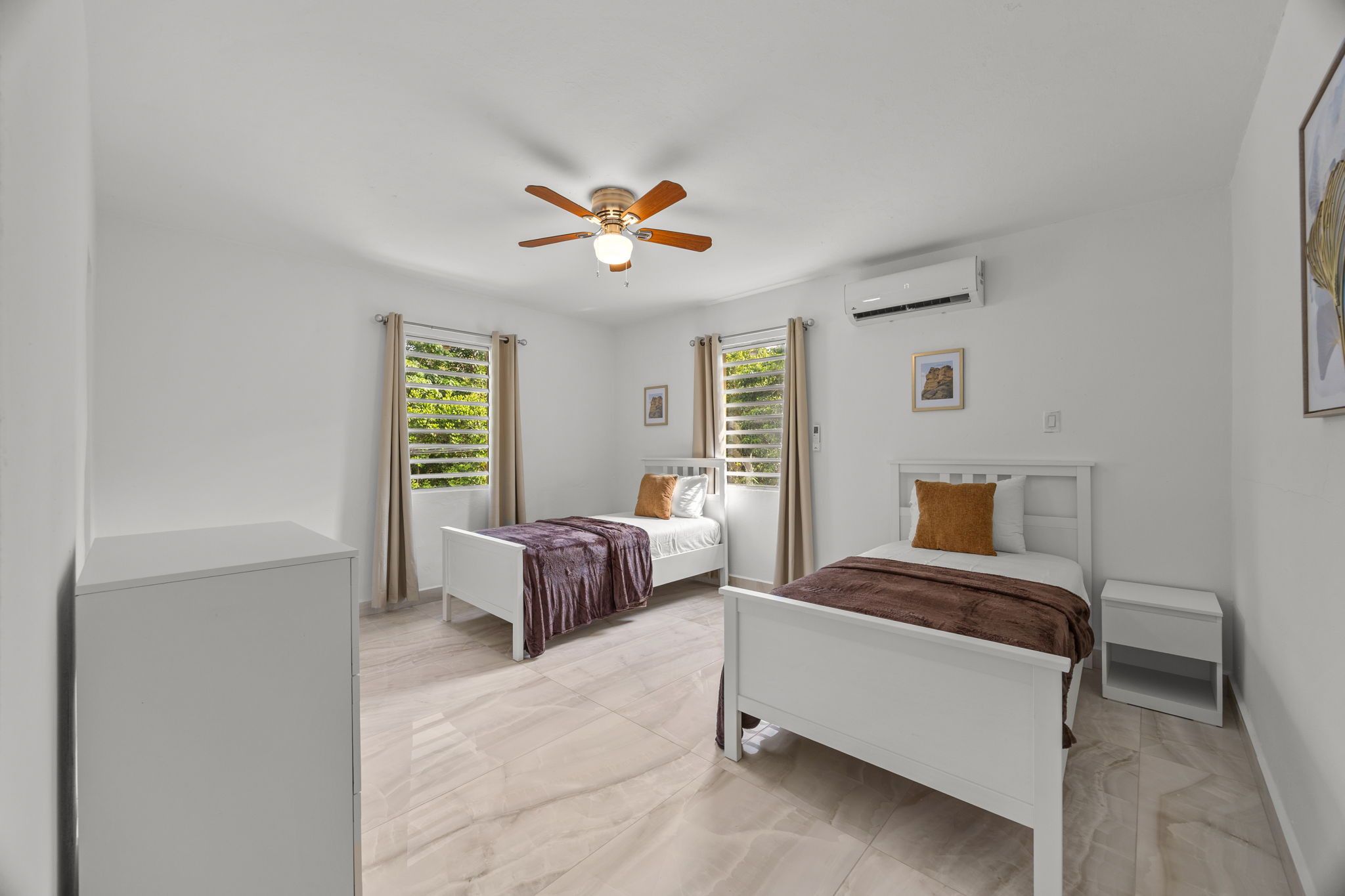 Quarto com duas camas, ventoinha no teto, aparelho de ar condicionado e janela com cortinas.