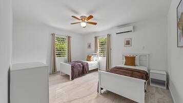 Quarto com duas camas, ventoinha no teto, aparelho de ar condicionado e janela com cortinas.