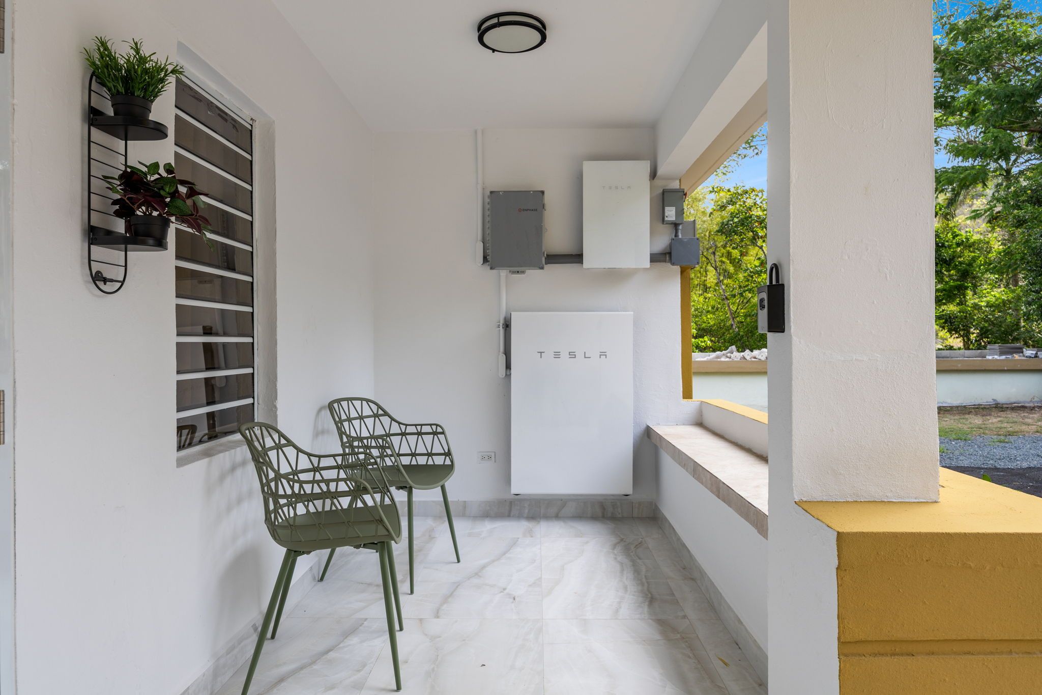 Uma pequena cozinha compacta com geladeira, pia e janela com vista para um jardim. Há duas cadeiras e um vaso de planta em uma prateleira.