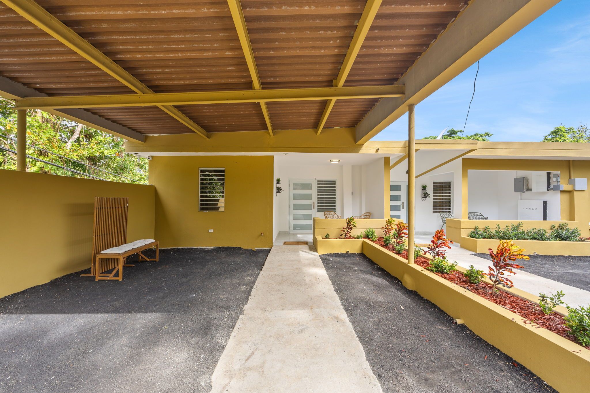 A covered outdoor area with a bench, a pathway, and a building with a Tesla charging station.