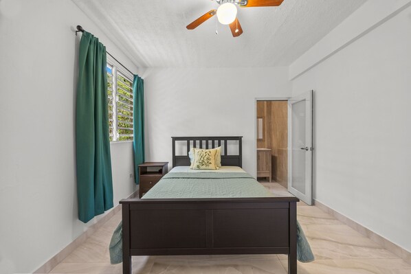 A bedroom with a bed, a nightstand, a ceiling fan, a window with curtains, and a door leading to a bathroom.