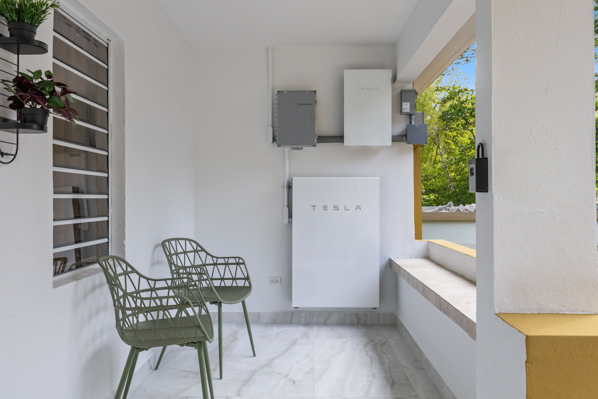 A small, modern kitchen with a white refrigerator, a built-in oven, and a wall-mounted microwave. There are two green chairs and a potted plant on the left.