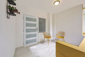 A hallway with a white door, two chairs, a small shelf with plants, and a ceiling light.