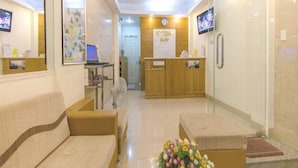 Un área de recepción con un sofá, una mesa baja con flores, un mostrador de recepción, un televisor y un reloj en la pared.