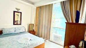 Habitación de hotel con una cama, un escritorio con una silla, un ventanal con cortinas, un cuadro en la pared y una lámpara en el techo.