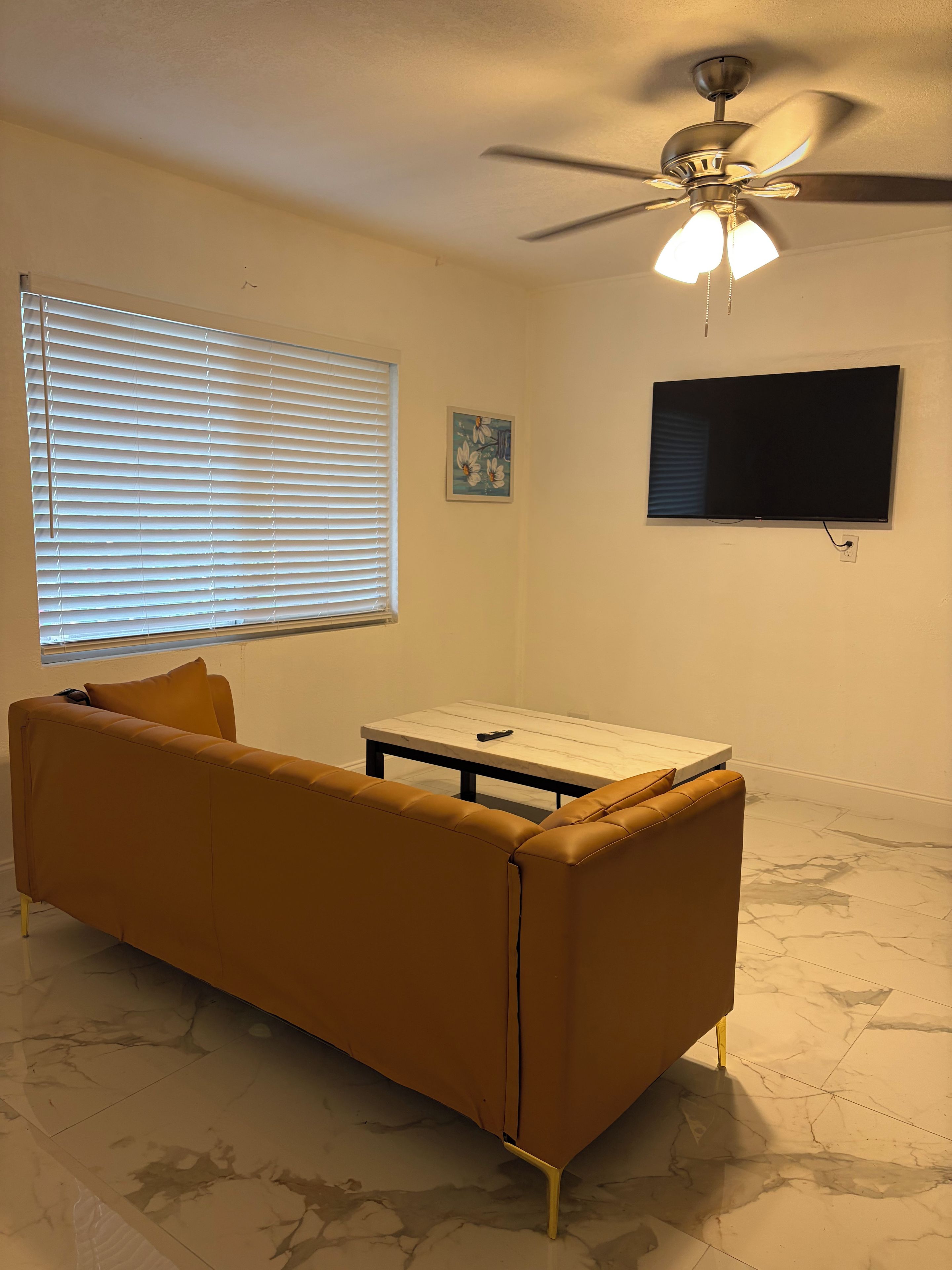 A living room with a sofa, a coffee table, a ceiling fan, a TV, and a window with blinds.