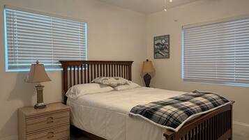 A bedroom with a bed, a nightstand, a lamp, a ceiling fan, and two windows with blinds.