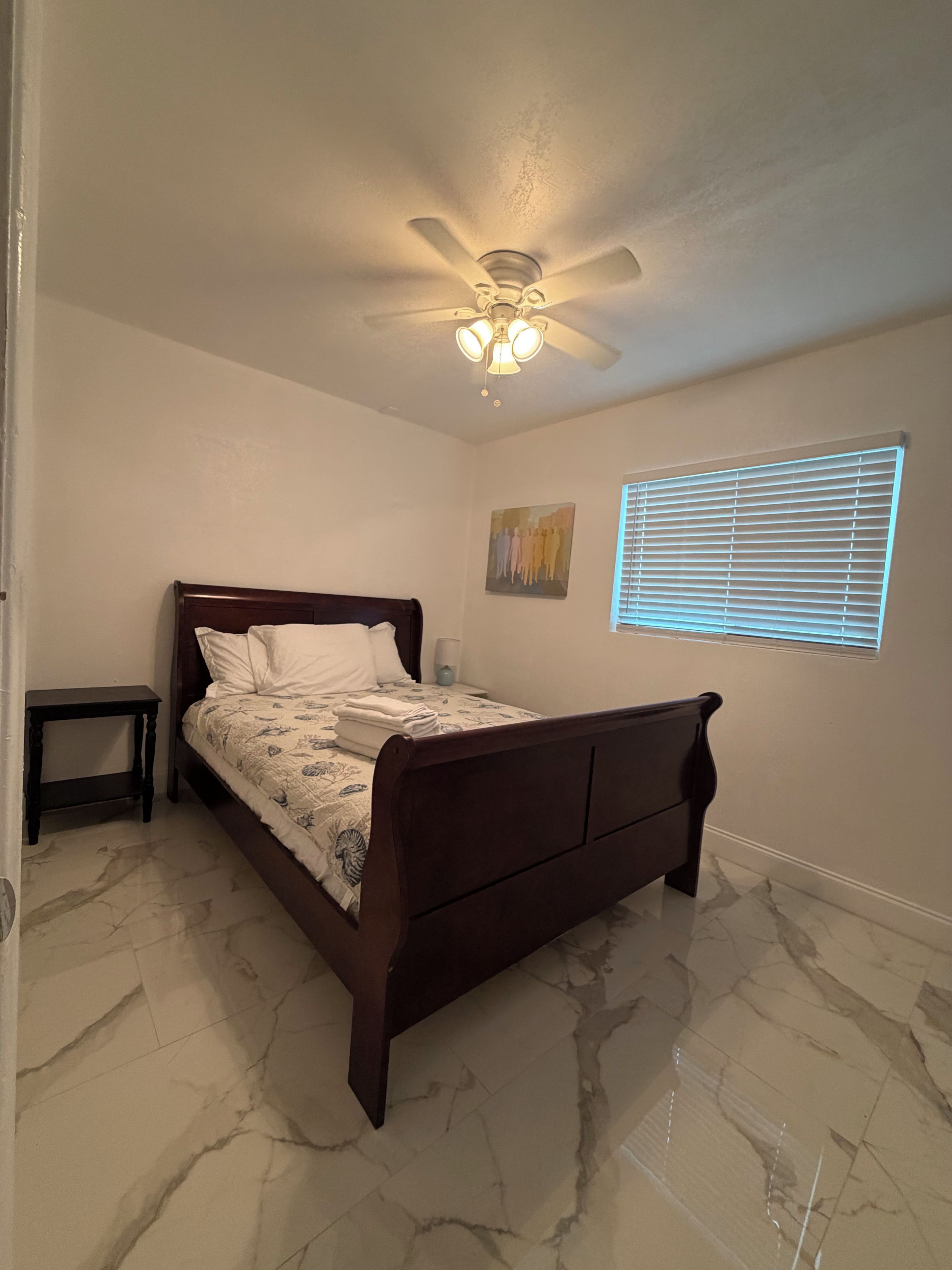 Une chambre à coucher avec un lit, un ventilateur de plafond, une fenêtre avec des stores et une petite table.