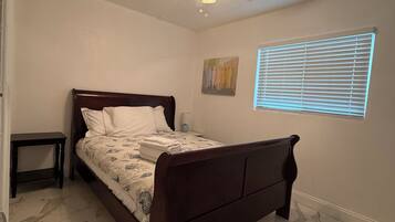 Une chambre à coucher avec un lit, un ventilateur de plafond, une fenêtre avec des stores et une petite table.