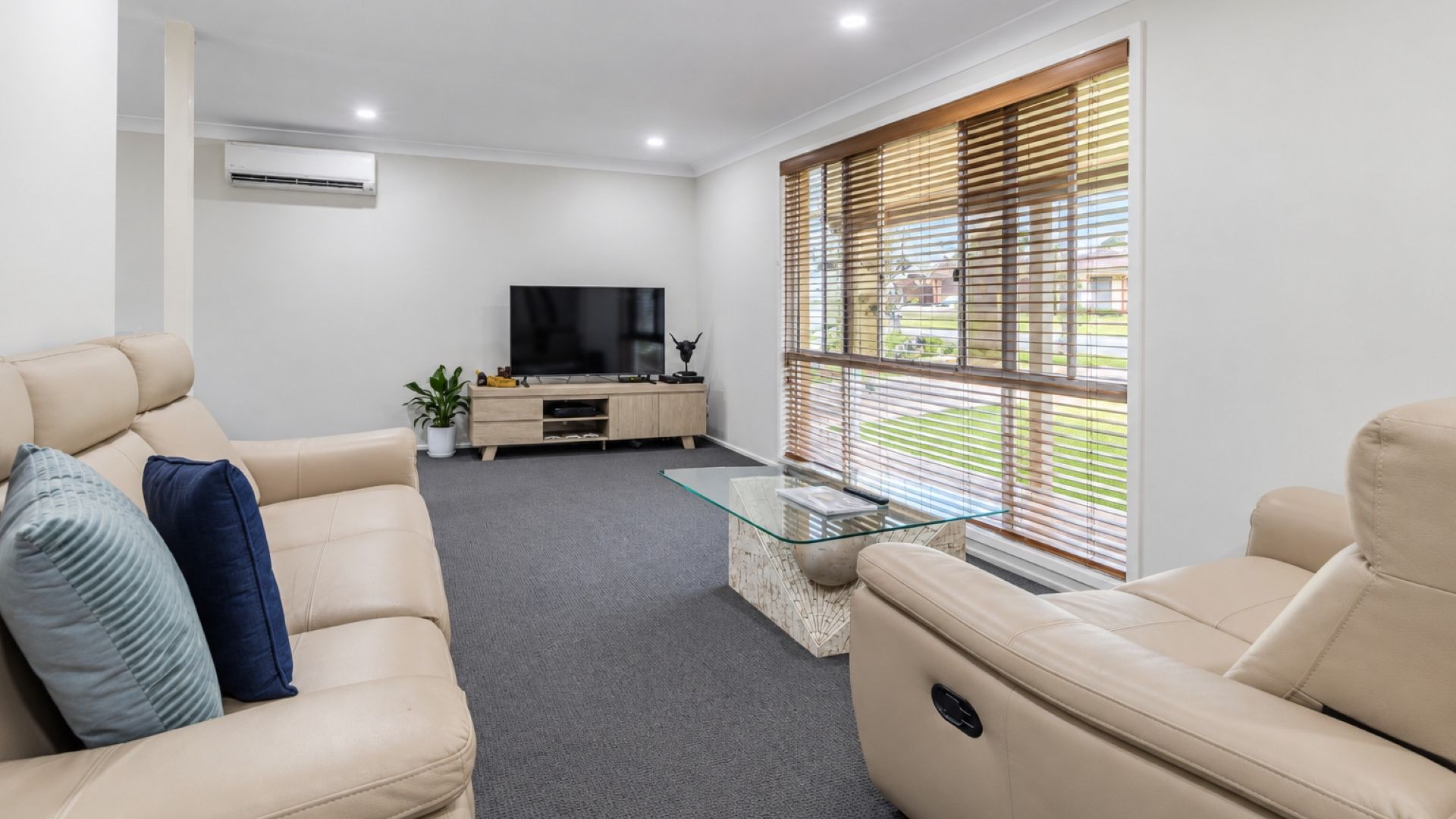 A modern living room with a sofa, a glass coffee table, a TV, and a window with blinds.