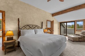 Ein Schlafzimmer mit einem großen Bett, einem Holz-Kopfteil, einem Nachttisch mit Lampe, einem Korbstuhl und Blick ins Freie durch eine Schiebetür.