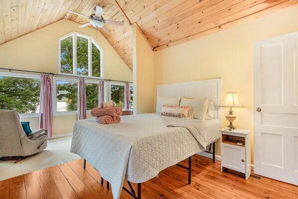 A bedroom with a bed, a nightstand, a chair, a window with curtains, and a ceiling fan.
