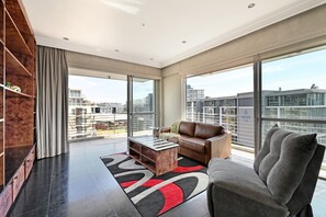 A spacious living room with a modern sofa, a coffee table, and a large window offering a city view. - Spacious Harbour Penthouse | Two Bedroom | Two Bathroom | Harbour & Mountain Views (Cape Town)