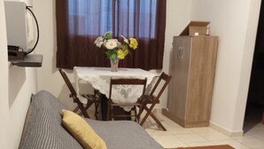 A single bed with a checkered blanket and a pillow, a small table with a vase of flowers, a wooden cabinet, and a window with curtains. - Apto Agua Marinha (Campos dos Goytacazes)
