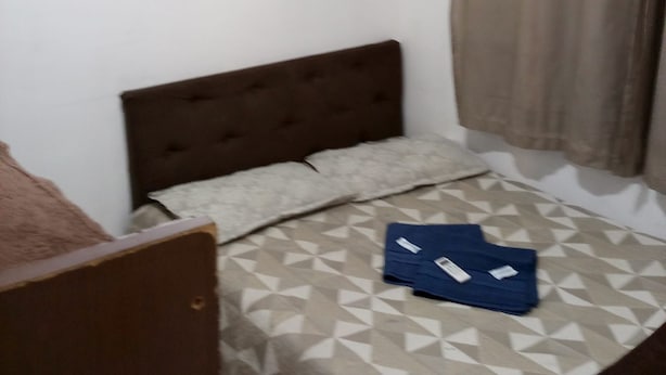 A small room with a bed, a pillow, a folded blue garment on the bed, a brown headboard, a white air conditioning unit on the wall, and a curtain. - Apto Agua Marinha (Campos dos Goytacazes)