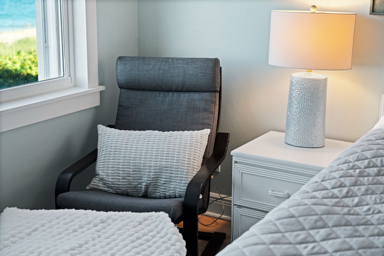 Un dormitorio con una silla, una mesita de noche con lámpara, una cama y una ventana con vistas a zonas verdes.