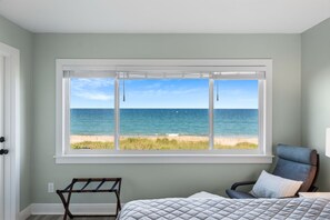 A bedroom with a large window overlooking the ocean, a bed with a quilted cover, and a chair with a pillow. - TIDE & SHELL - Remodeled Oceanfront Jensen Beach Retreat w/Private Beach & Pools (Jensen Beach)