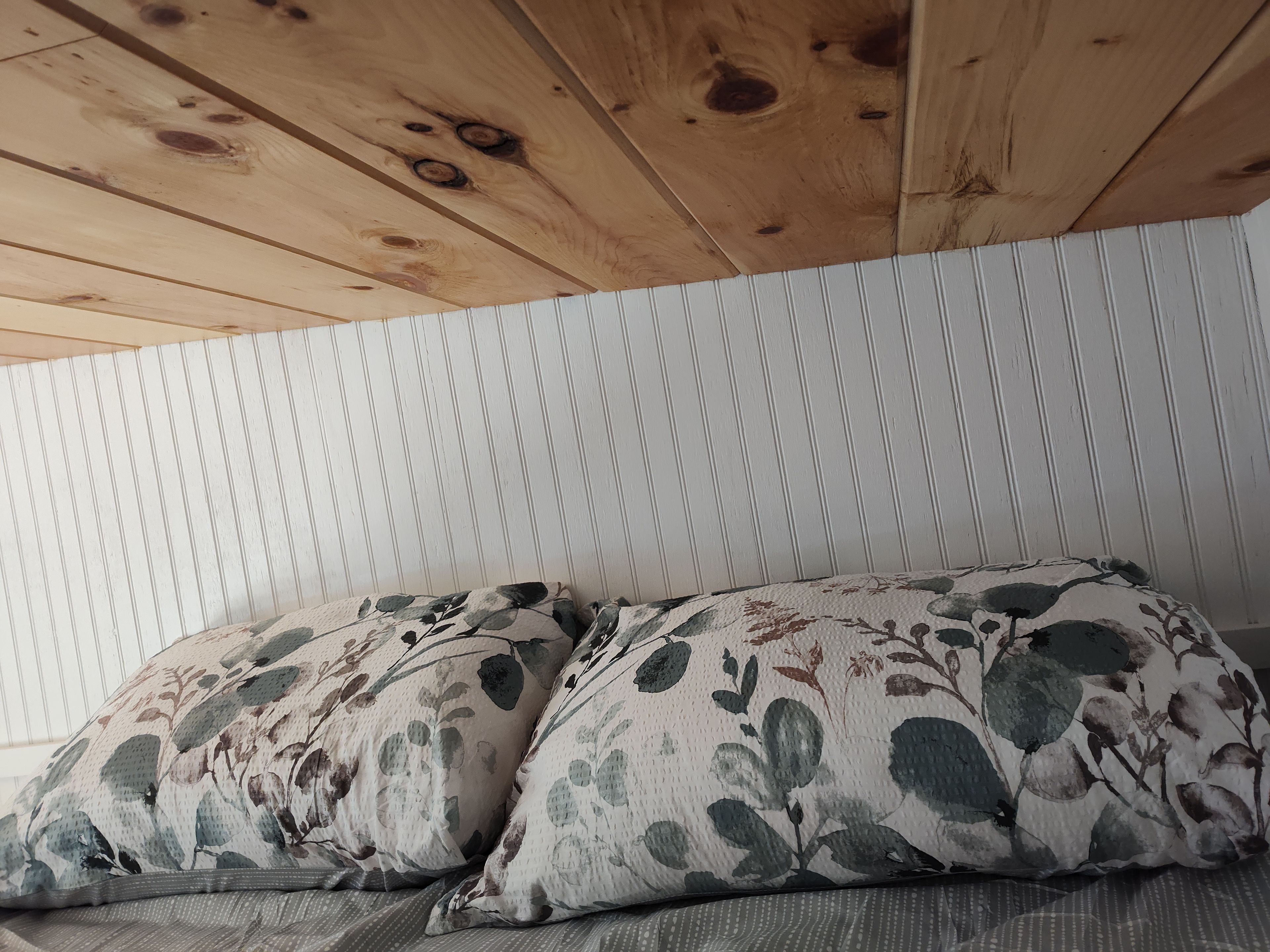 Une pièce avec un plafond en bois, un lit agrémenté d’oreillers à motifs floraux et un mur à rayures blanches.