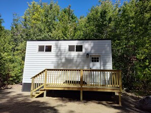 A single-story cabin with a wooden deck and a small porch, surrounded by dense forest.
