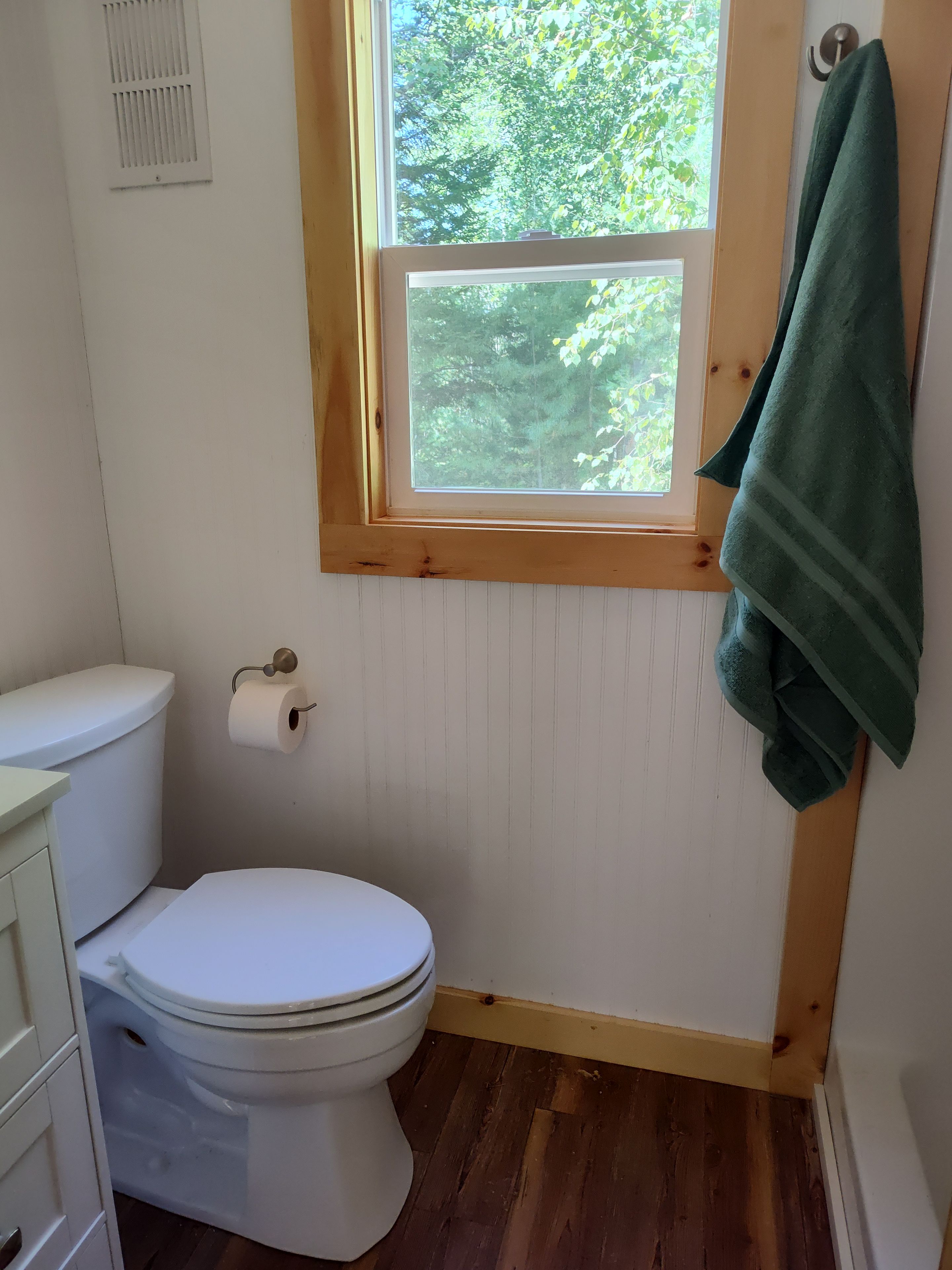 A bathroom with a toilet, a window with a view of trees, and a towel hanging on the wall.
