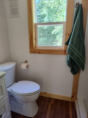 A bathroom with a toilet, a window with a view of trees, and a towel hanging on the wall.