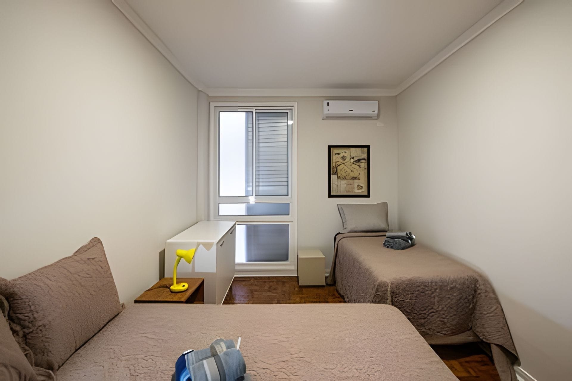A small, well-lit room with two beds, a desk, a chair, and a window with blinds.