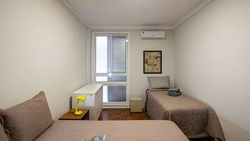A small, well-lit room with two beds, a desk, a chair, and a window with blinds.