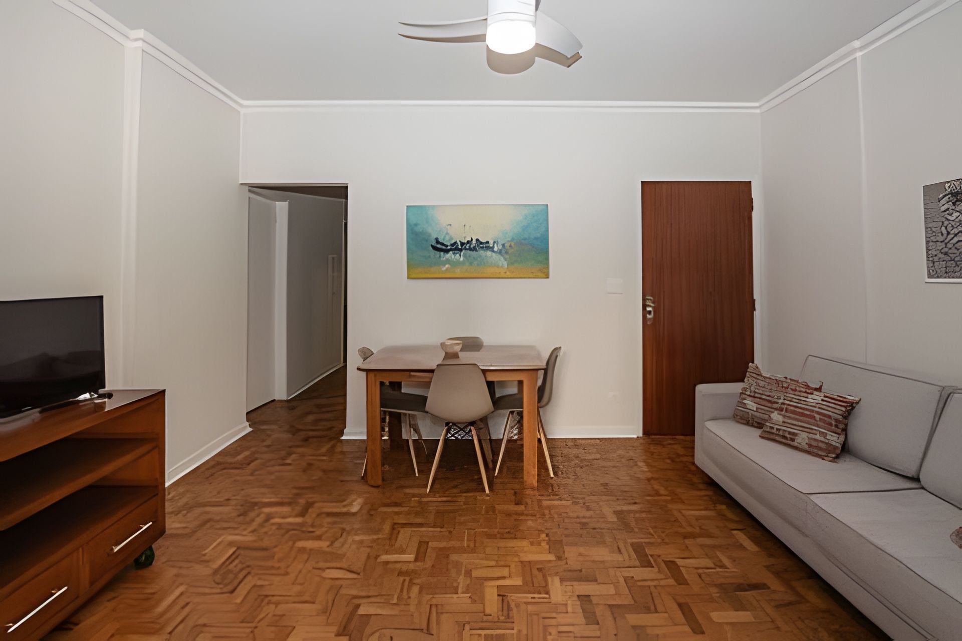Uma sala de estar com piso de madeira em padrão herringbone, uma mesa de jantar com cadeiras, um sofá, uma televisão sobre um móvel de madeira e um ventilador de teto.