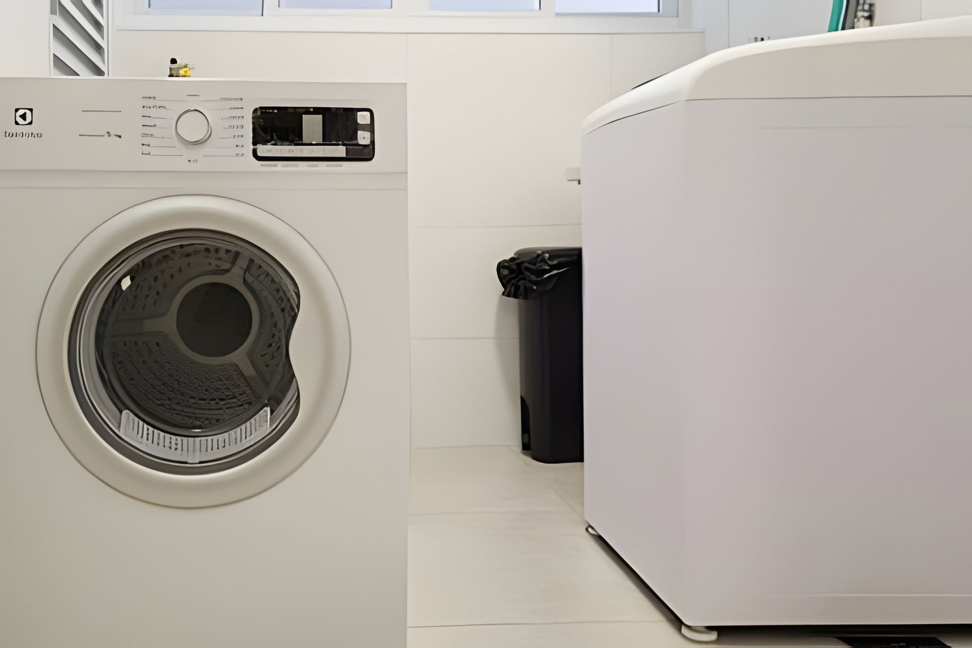 A front-loading washing machine and dryer in a laundry room with a trash can nearby.