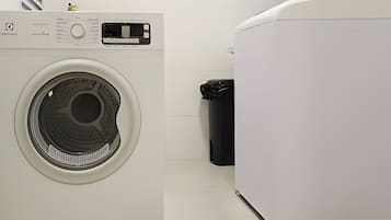 A front-loading washing machine and dryer in a laundry room with a trash can nearby.