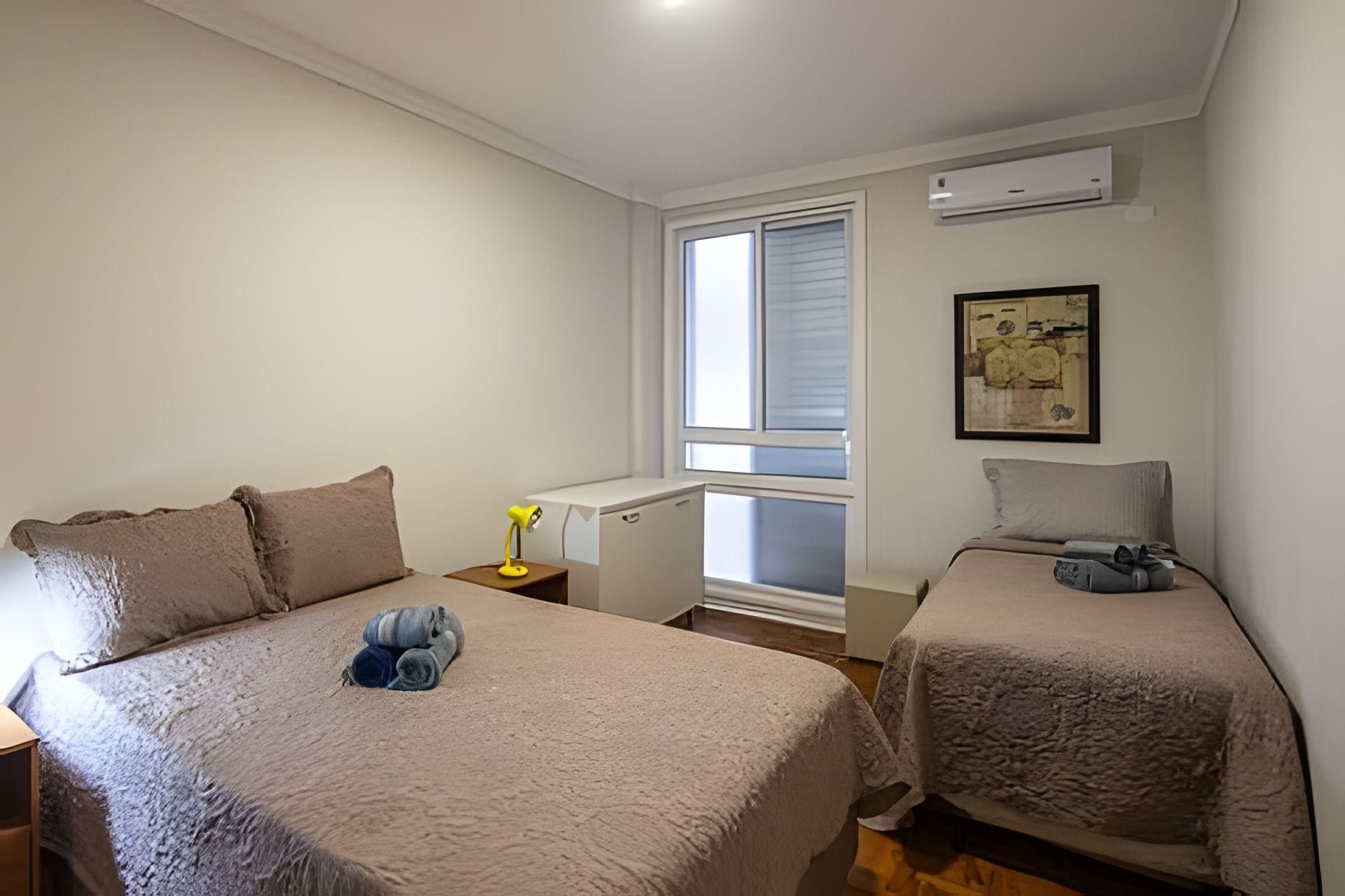 Un dormitorio con dos camas, una ventana con persianas, aire acondicionado y una mesita de noche con lámpara.