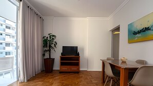 Uma sala de estar moderna com piso de madeira em padrão herringbone, uma janela grande com cortinas, um vaso de planta, uma TV de tela plana em um suporte de madeira, uma mesa de jantar com cadeiras e um quadro na parede.