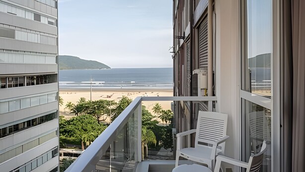 Uma varanda com vista para a praia e o oceano, cadeiras brancas e guarda-corpo de vidro.
