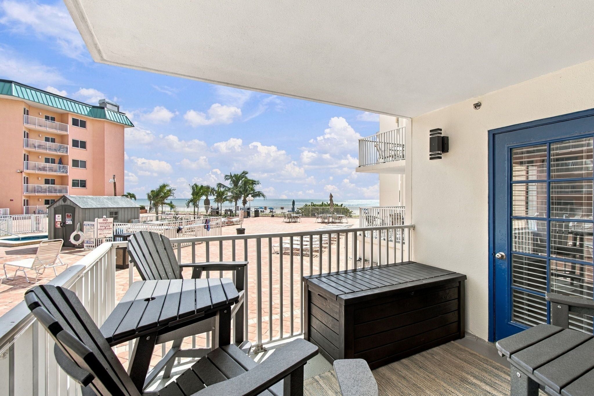 En balkon med udendørs møbler, udsigt til stranden og en bygning i baggrunden.