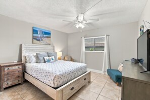 A bedroom with a bed, a nightstand, a dresser, a ceiling fan, a painting, and a window with a view. - Coastal Ocean View Condo at Beach Palms (Indian Shores)