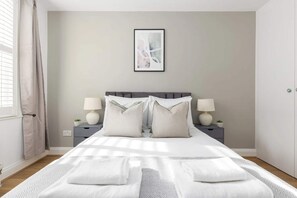 A neatly made bed with white linens and pillows, two bedside lamps, a framed abstract artwork on the wall, and a window with curtains. - 2 Bed Private Garden near Shepherds Bush Westfield (London)
