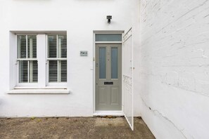 Ein weißes Gebäude mit einer Tür und einem Fenster, ein kleines Schild über der Tür und eine Ziegelwand zur rechten Seite.