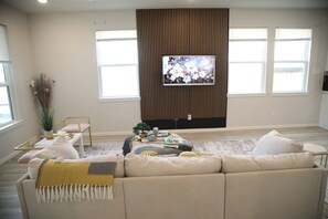 A living room with a beige sofa, a coffee table, a TV mounted on a wooden wall, and a window with blinds.