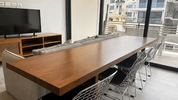 Un espace repas moderne avec une longue table en bois, des chaises en métal, un téléviseur posé sur un meuble et une vue sur les bâtiments à travers de grandes fenêtres.