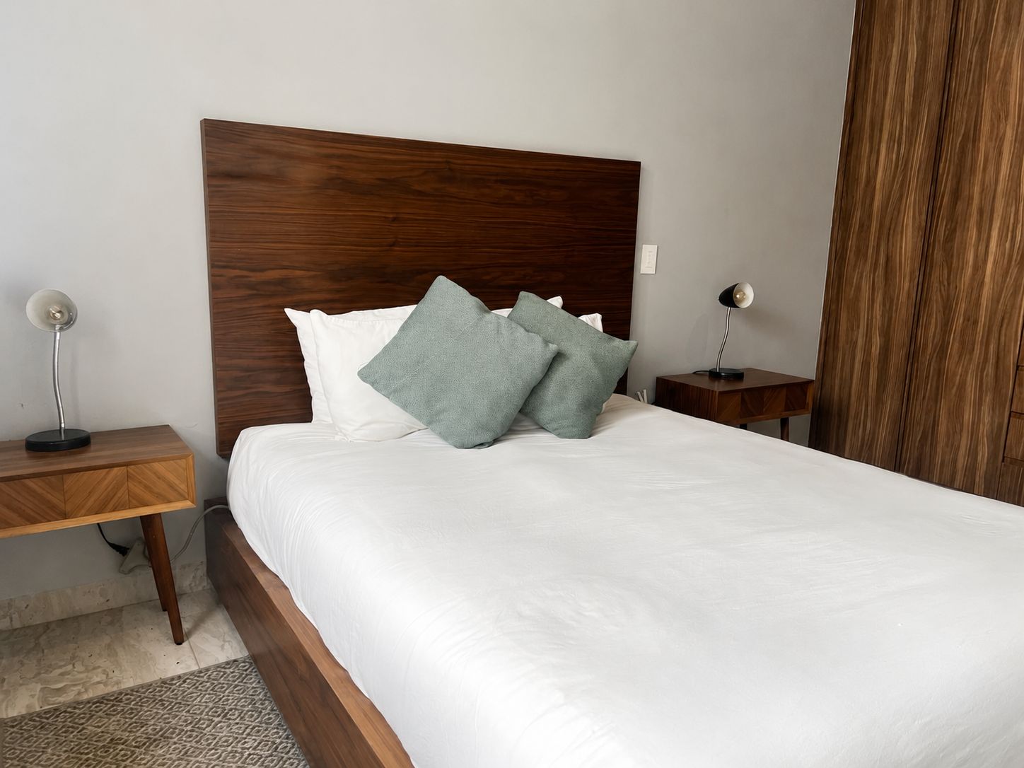 Un lit bien fait, avec une tête de lit en bois, deux tables de chevet avec des lampes et un tapis à motifs sur le sol.