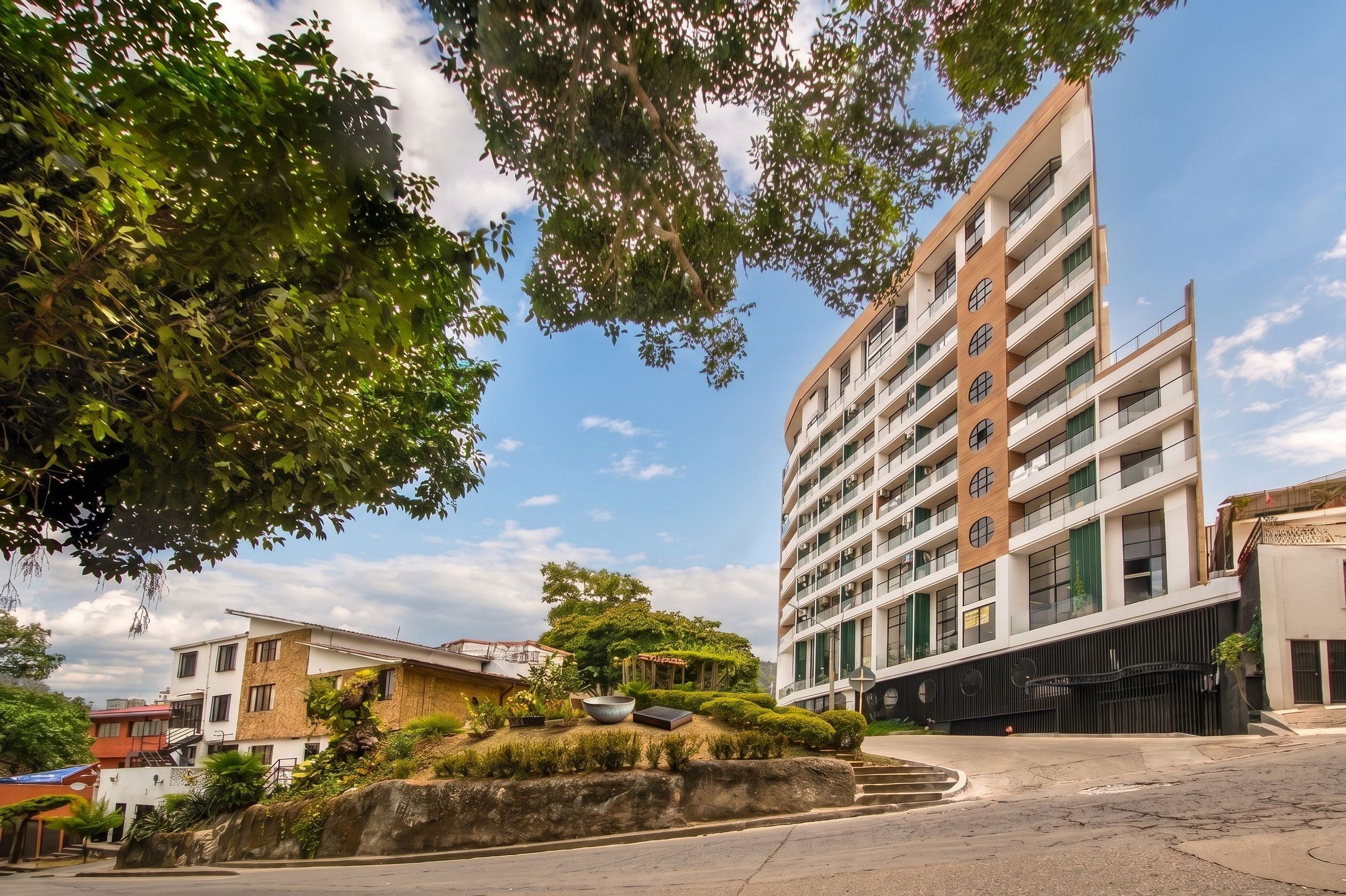 Un edificio residencial de varios pisos con balcones, una calle pavimentada y zonas verdes circundantes.