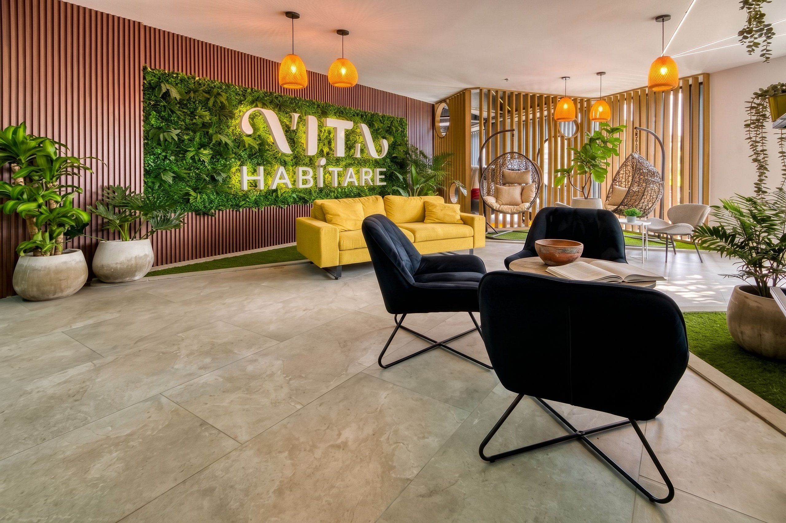 Uma sala de estar moderna com um sofá amarelo, cadeiras pretas e uma parede verde com o texto 'VITA HABITATRE'.