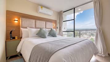 A hotel room with a large bed, a nightstand, an air conditioning unit, a view of the city through a large window, and a balcony.