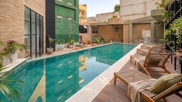 A swimming pool with lounge chairs and potted plants, surrounded by buildings with large windows and a brick facade.