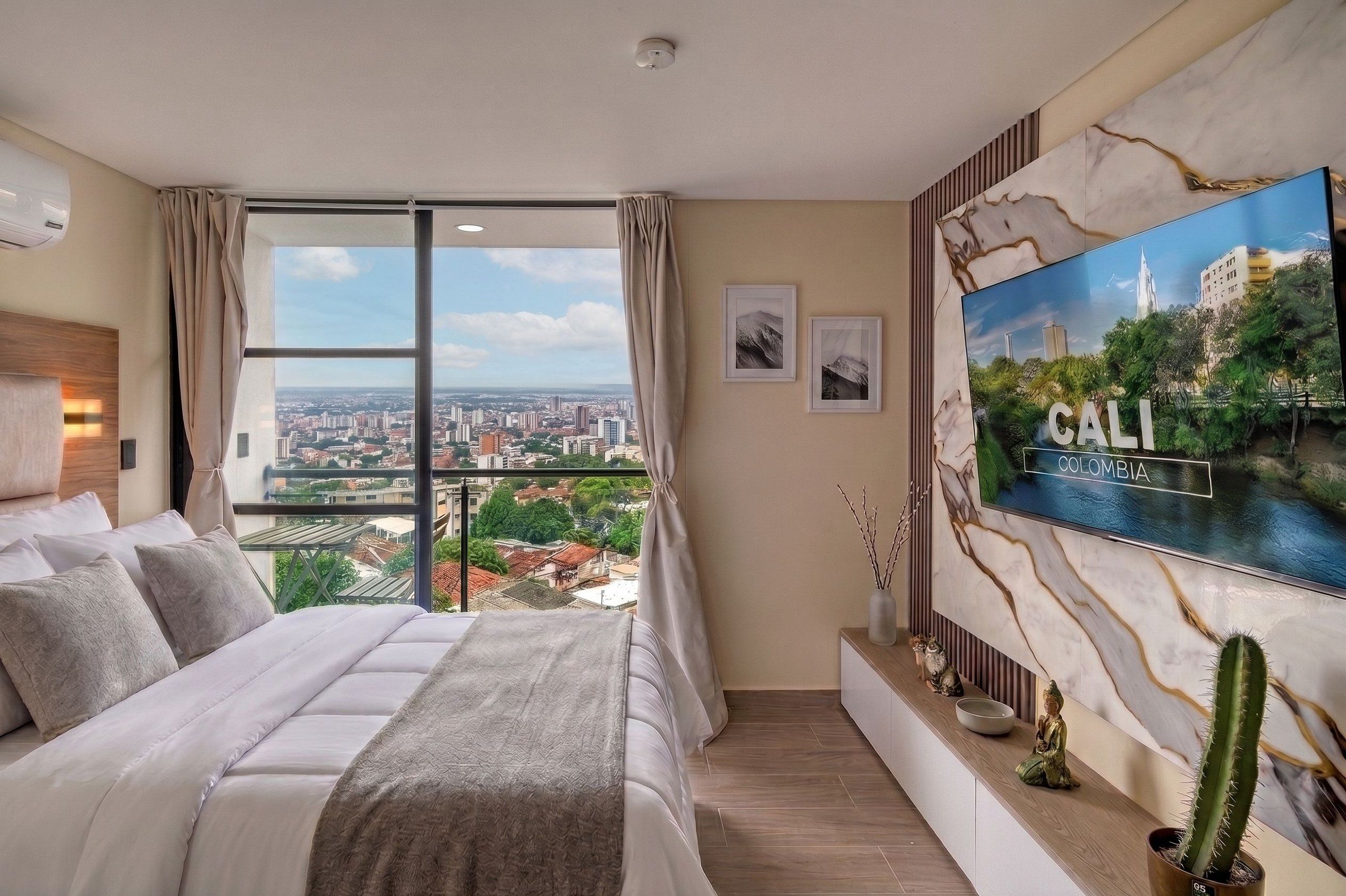 Habitación de hotel con una cama grande, vista a la ciudad por la ventana, un televisor de pantalla plana montado en la pared y un jarrón decorativo con un cactus.