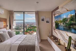 Habitación de hotel con una cama grande, vista a la ciudad por la ventana, un televisor de pantalla plana montado en la pared y un jarrón decorativo con un cactus.