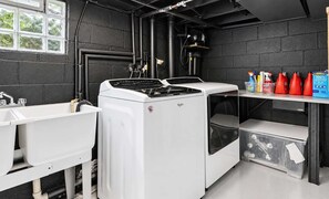 A laundry room with a front-loading washer and dryer, a sink, and a shelf with cleaning supplies. - Riverside Retreat | Hot Tub • Sauna • Private Dock (Saugatuck)
