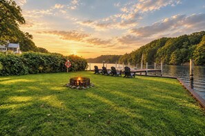 Ein Seeufer mit einer Feuerstelle und Stühlen, umgeben von üppiger Vegetation und Blick auf den Sonnenuntergang.